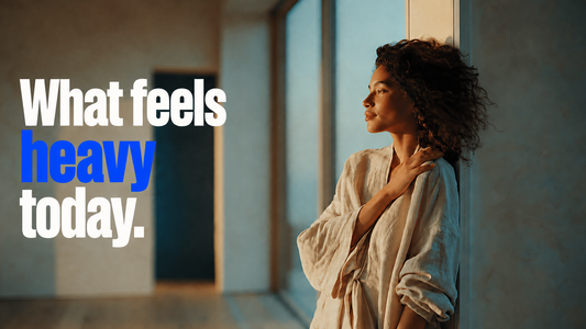 What feels heavy today — a young woman with curly hair leans against a sunlit doorway in a quiet interior, looking out a window in soft afternoon light, holding her shoulder in a still moment of carrying.