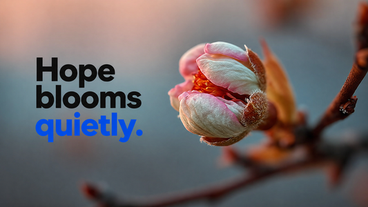 Hope blooms quietly — a single pale pink and cream blossom opens on a bare branch against a soft slate-blue and peach background, captured in close intimate focus.