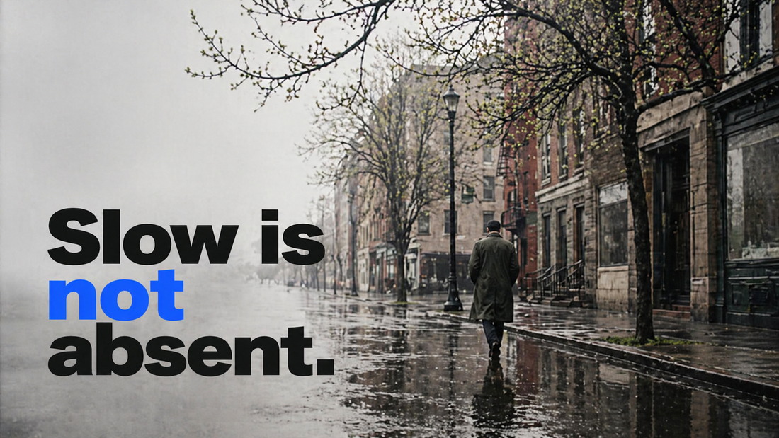 Slow is not absent — a man in a dark coat walks alone down a misty rain-slick city street lined with bare trees just beginning to bud into spring green, soft fog blurring the distance.